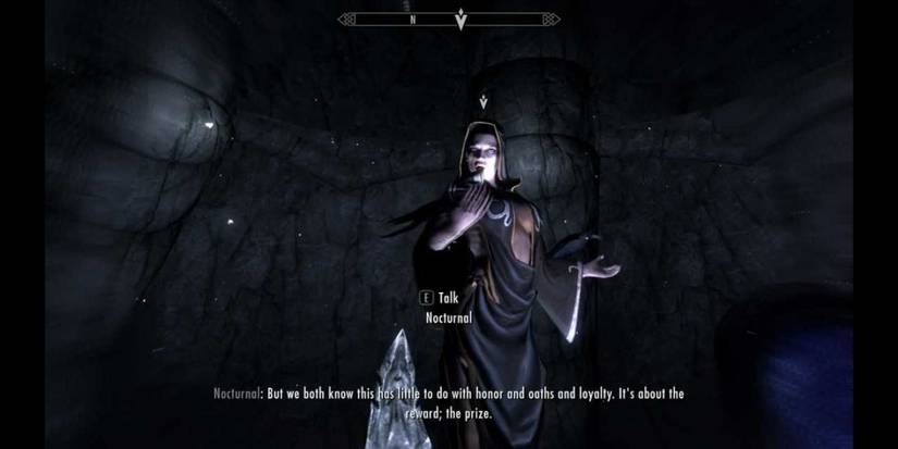 A levitating woman dressed in loose black robes looks down as she speaks at the viewer.