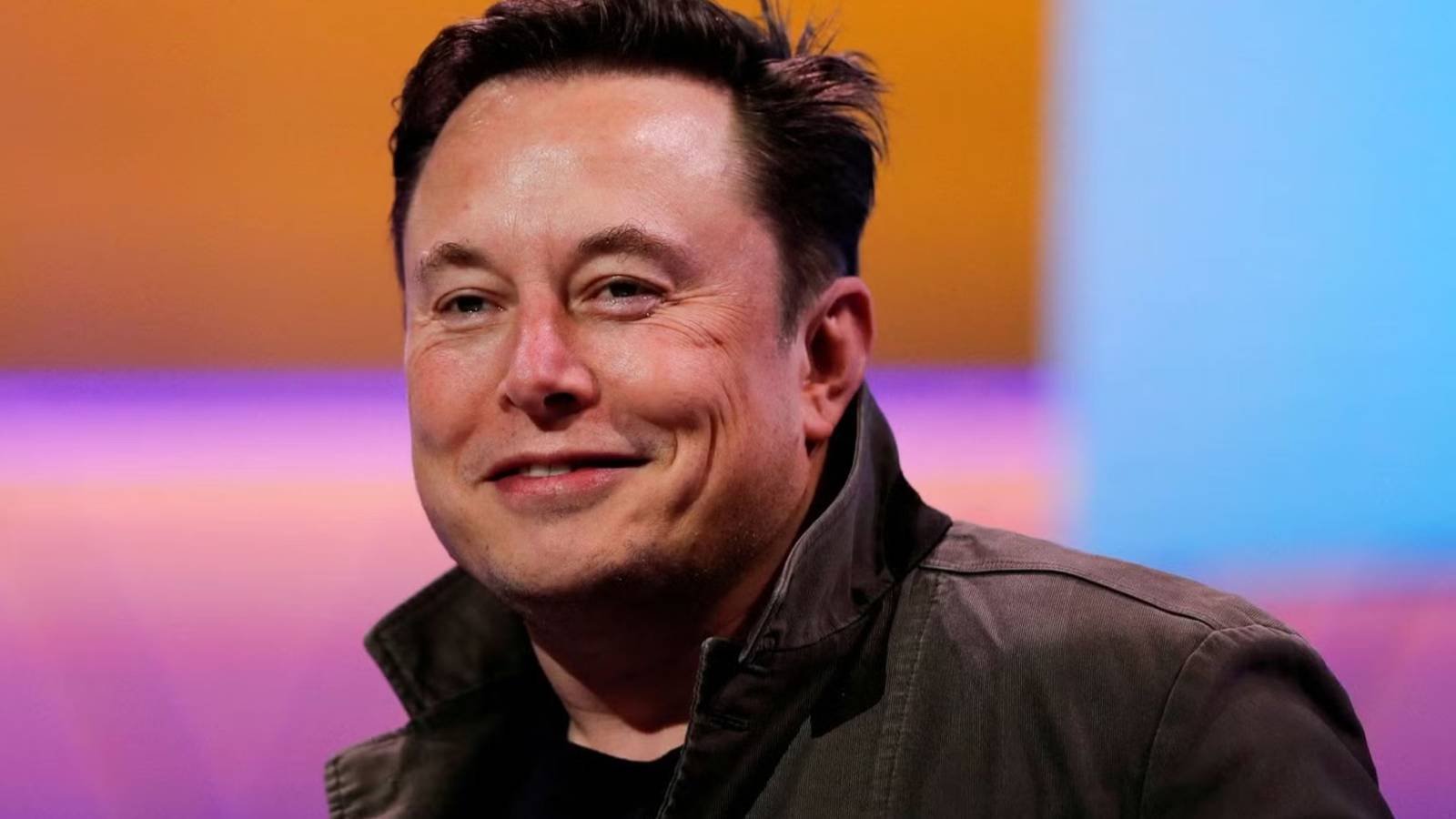 Elon Musk smiling. There is a colourful background.