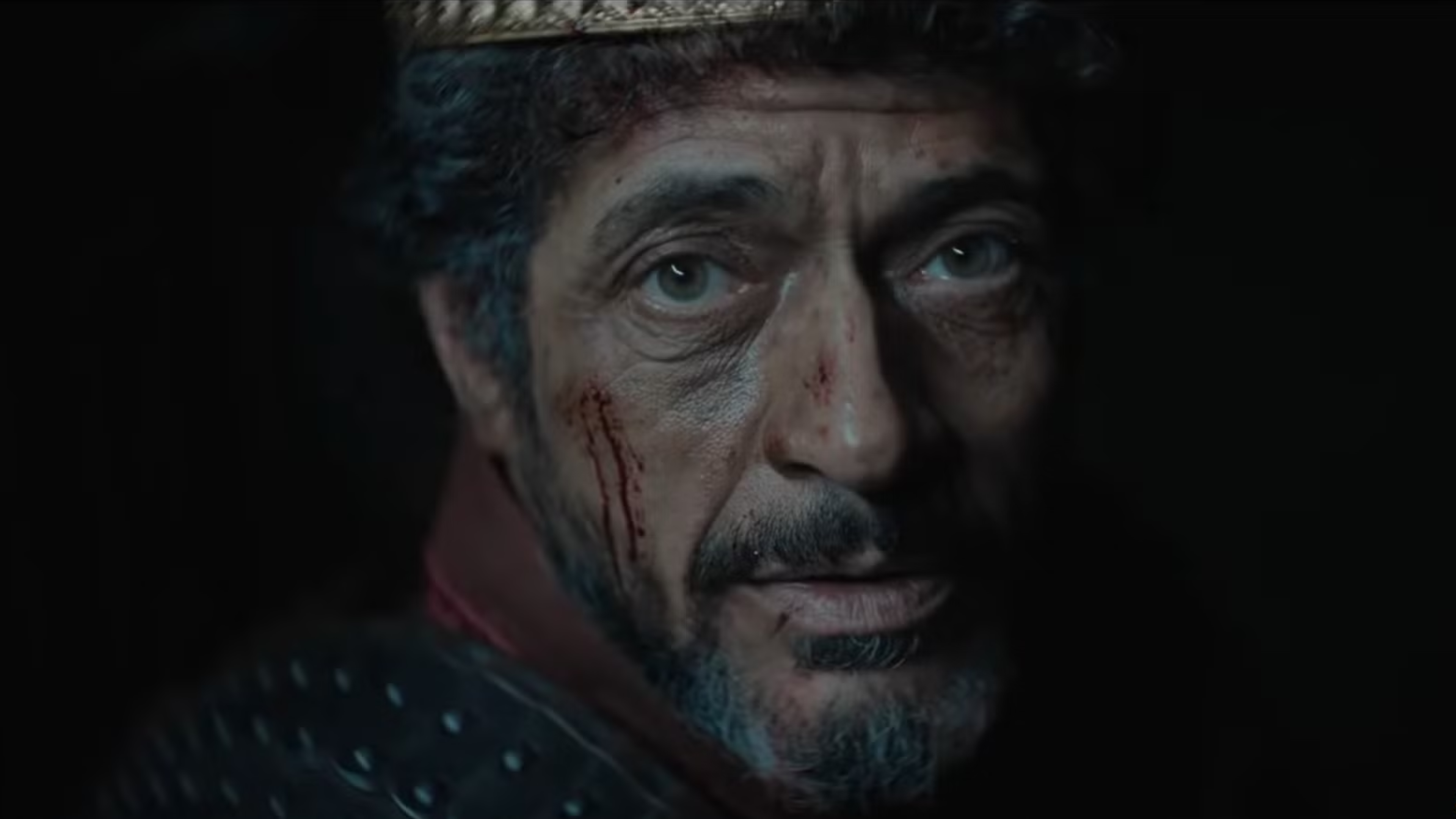 A close up of a middle aged man's face. He has blood on his face, greying hair, and appears to be wearing a crown.