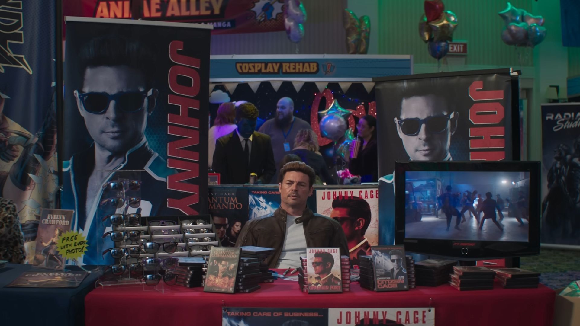 A still from Mortal Kombat 2, showing Johnny Cage sitting at an empty comic con stall, surrounded by his merch.-1