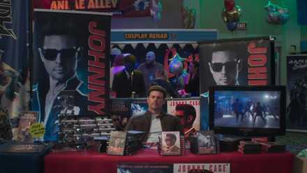 A scene from Mortal Kombat 2. It shows Johnny Cage sitting in an empty Comic Con booth surrounded by his merchandise.-1