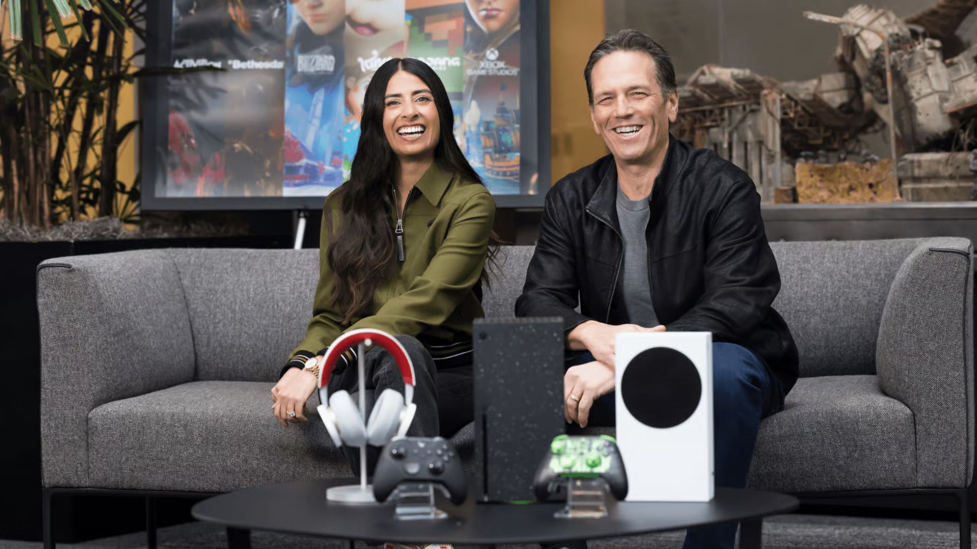 Asha Sharma and Phil Spencer sitting on a sofa with Xbox models in front of them.-1
