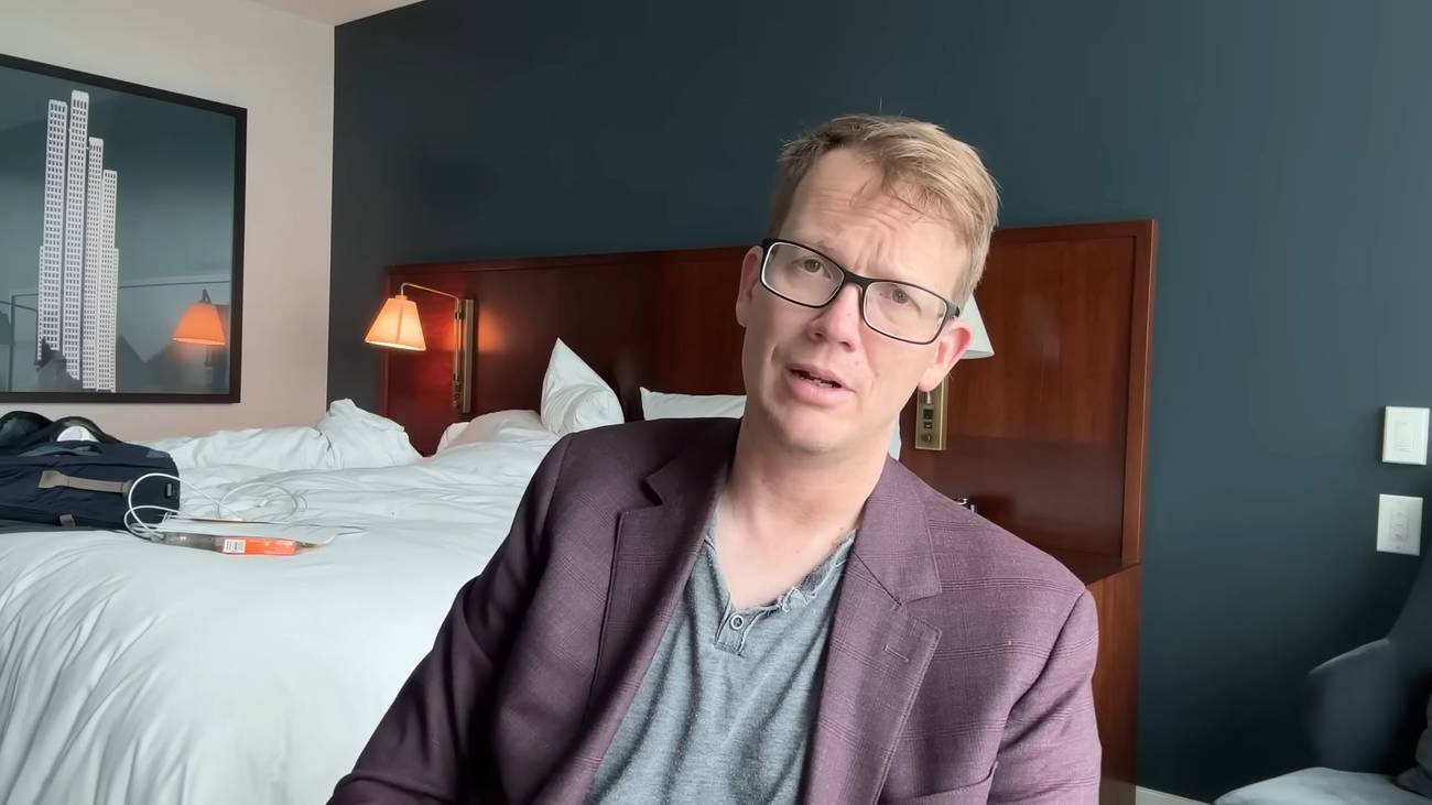 Hank Green in a hotel room, sitting in front of his bed.