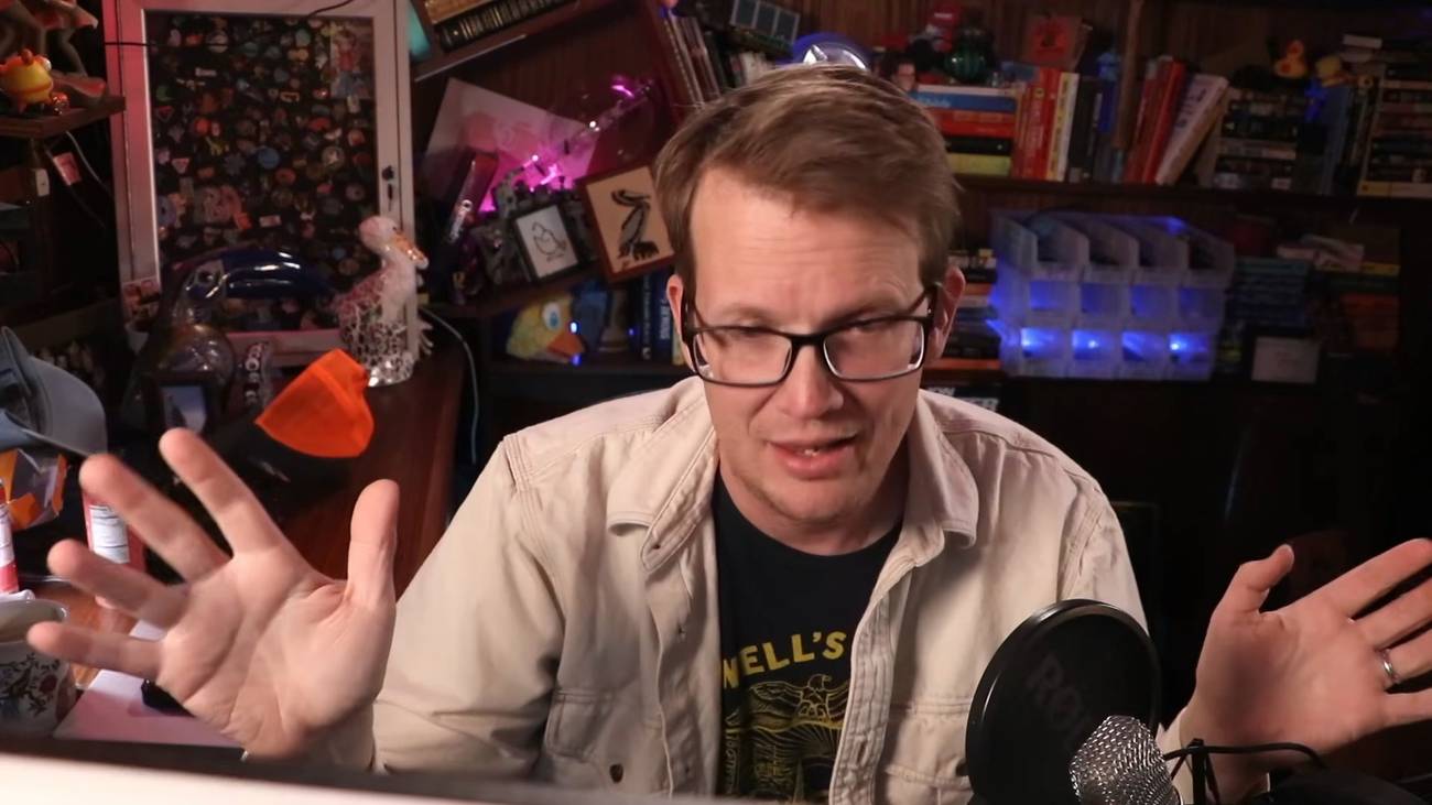 Hank Green putting up his hands at his desk in front of a mic.