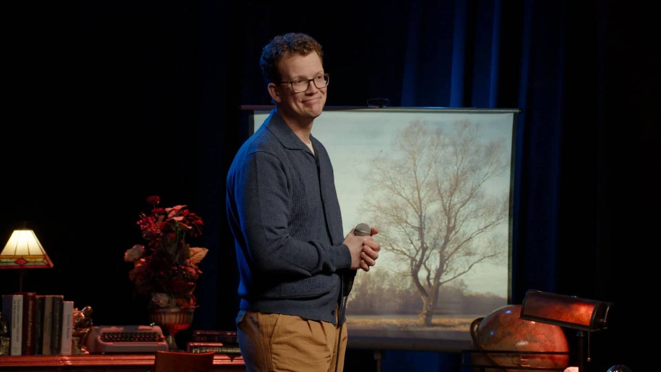 Hank Green standing in front of a painting of a tree in his Dropout comedy special.