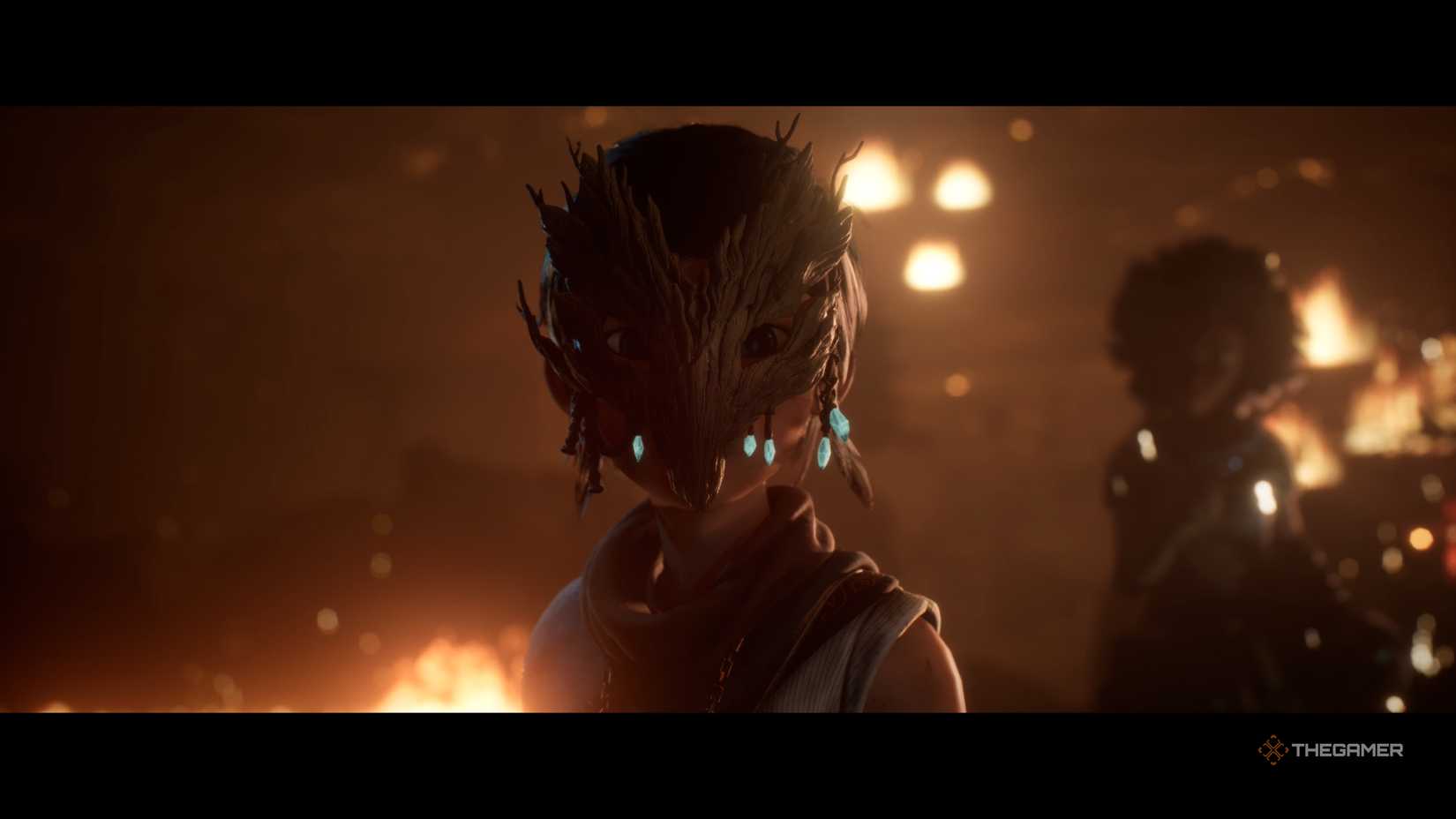In Kena: Bridge of Spirits, Kena is seen wearing a wooden mask while a fire burns, with another character standing behind her.